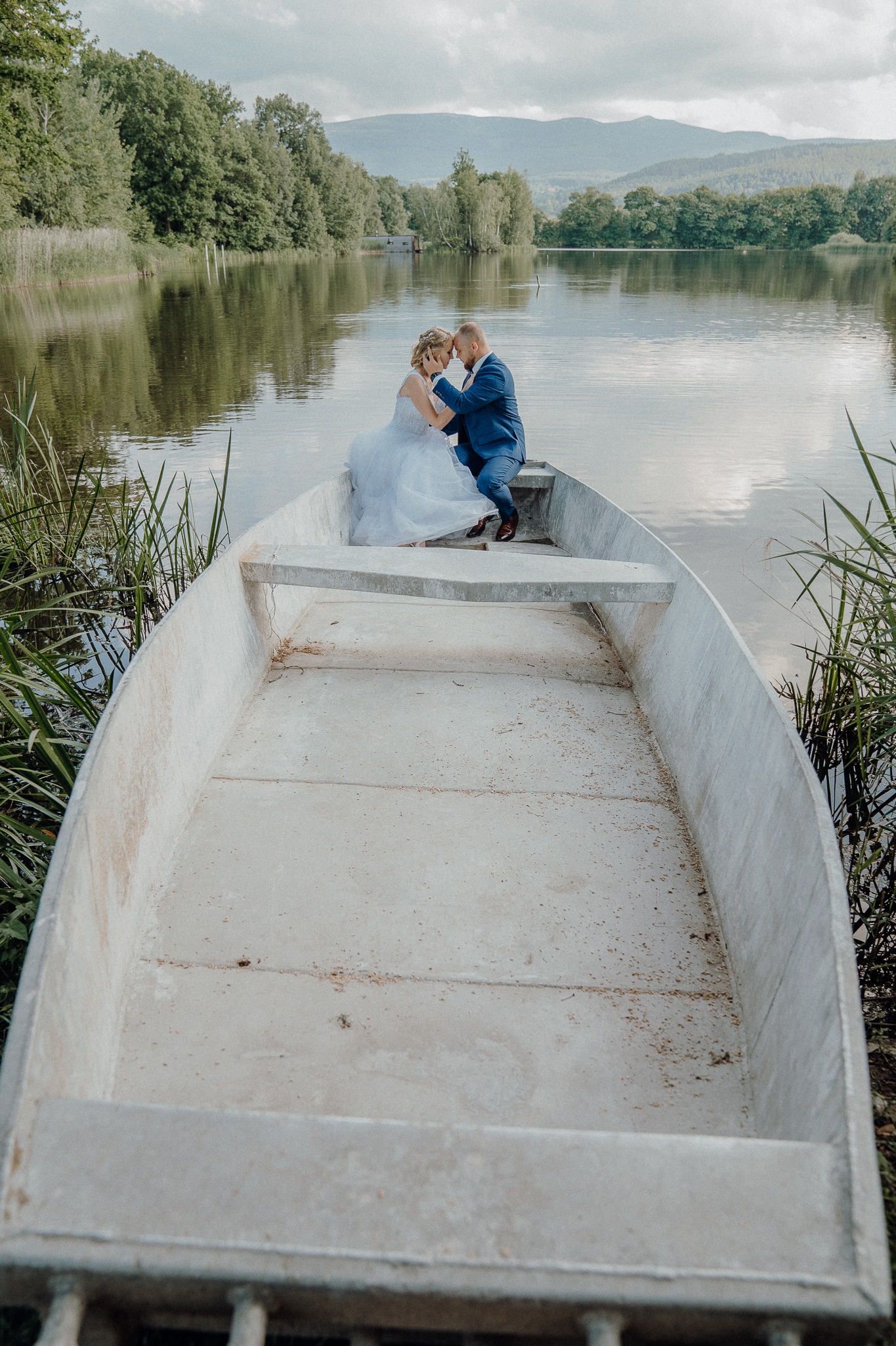 Para młoda siedząca w łodzi na jeziorze, mężczyzna w niebieskim garniturze, kobieta w białej sukni ślubnej, drzewa i góry w tle.