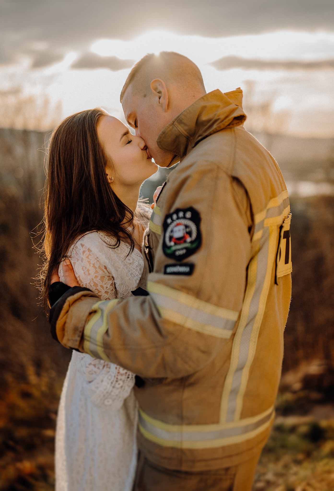 Zakochana para: kobieta w koronkowej sukience i mężczyzna w stroju strażaka, pocałunek na tle zachodzącego słońca.