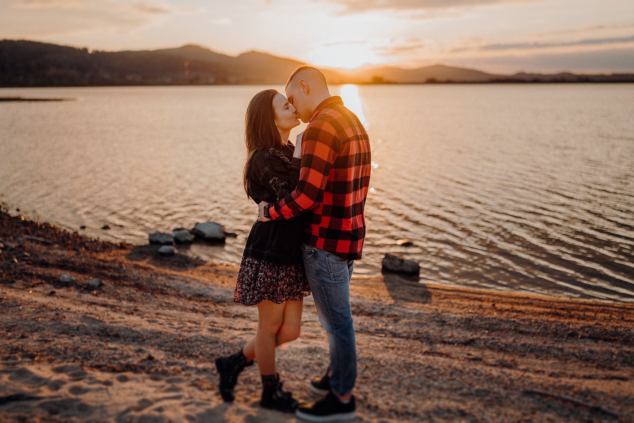 Para całująca się na brzegu jeziora o zachodzie słońca, kobieta w kwiecistej sukience i czarnej kurtce, mężczyzna w czerwonej koszuli w kratę.