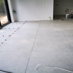 Magwest - Jasnoszara podłoga z płytek w trakcie układania, z systemem poziomowania na krawędziach przy oknie i widocznymi narzędziami oraz wiadrami w tle.