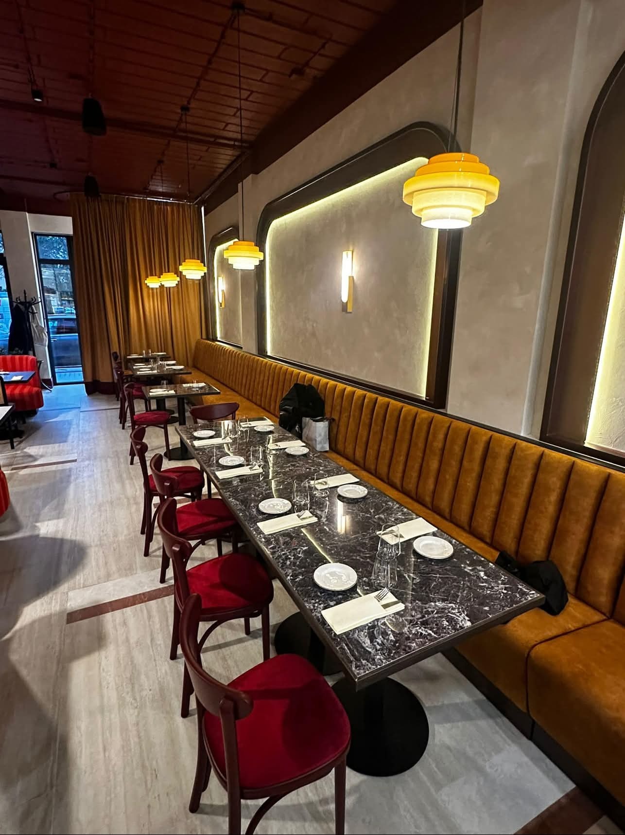 Wnętrze restauracji z długim, marmurkowym stołem, czerwonymi krzesłami i pomarańczową kanapą wzdłuż ściany, oświetlone żółtymi lampami w stylu retro i ozdobnymi kinkietami.