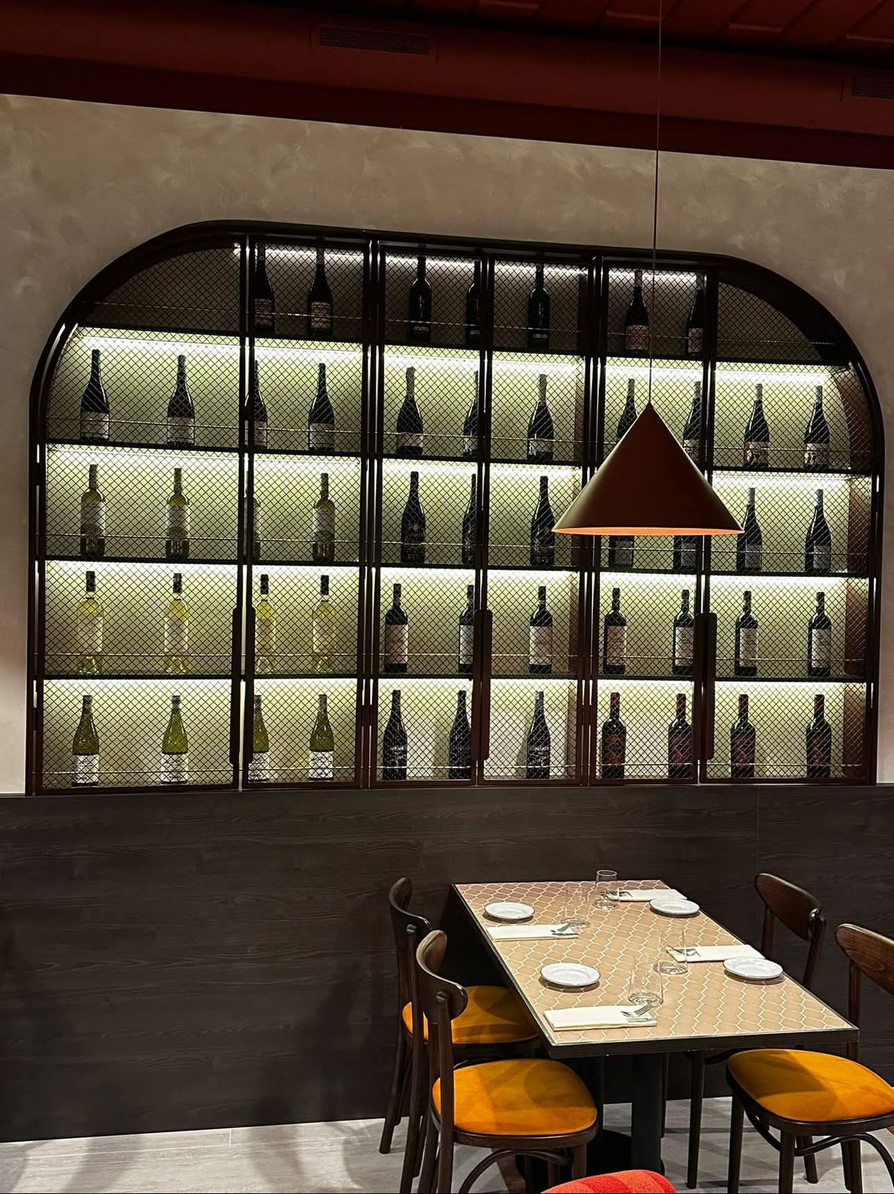 Aranżacja wnętrza restauracji z podświetlaną półką na wina z metalowej siatki, stołem z nakryciem i krzesłami z pomarańczowymi siedziskami, oświetlona wiszącą lampą z brązowym kloszem.