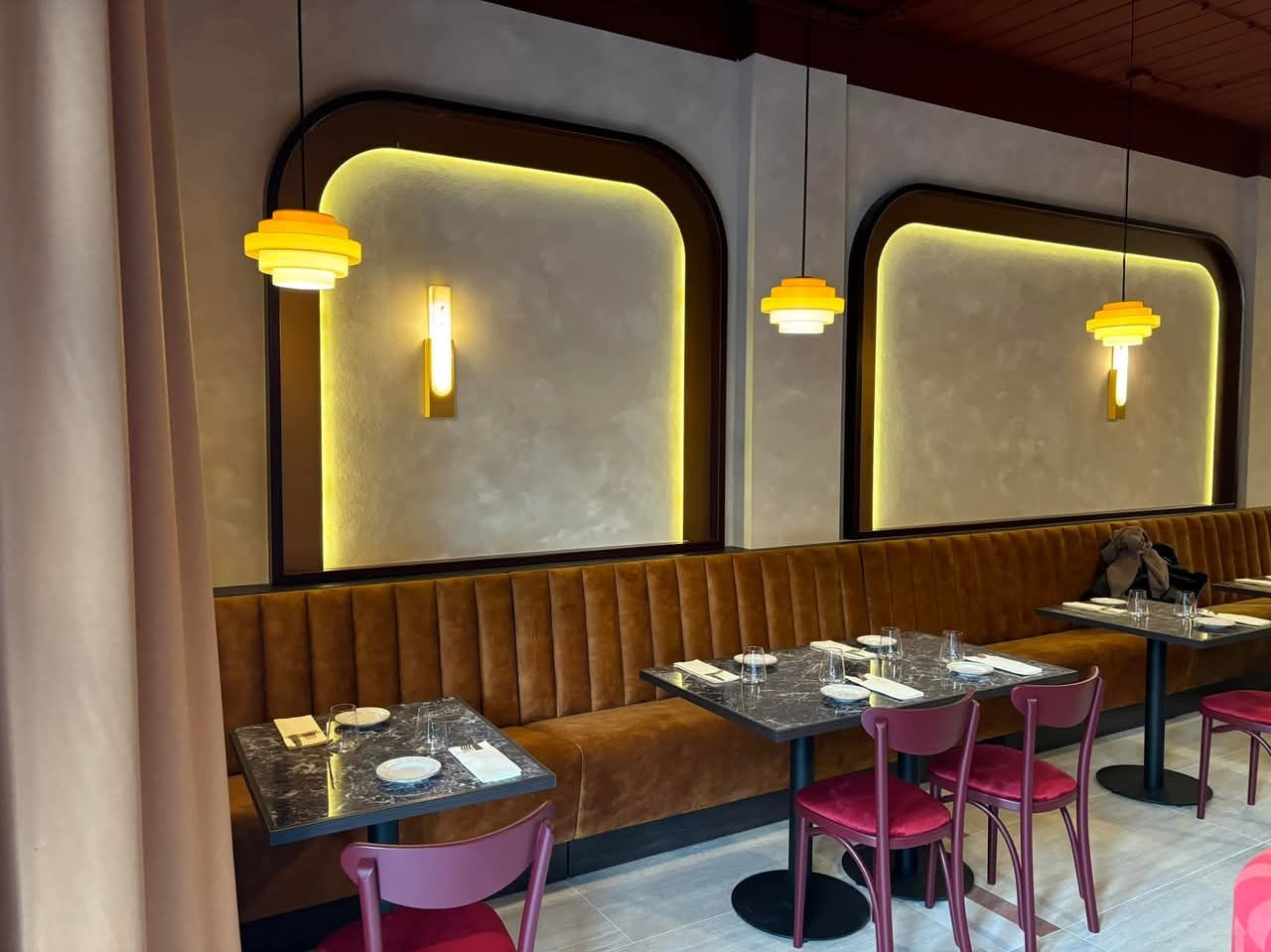 Wnętrze restauracji z brązową, pluszową kanapą, marmurowymi stolikami, bordowymi krzesłami i dekoracyjnymi, podświetlanymi wnękami w ścianie.