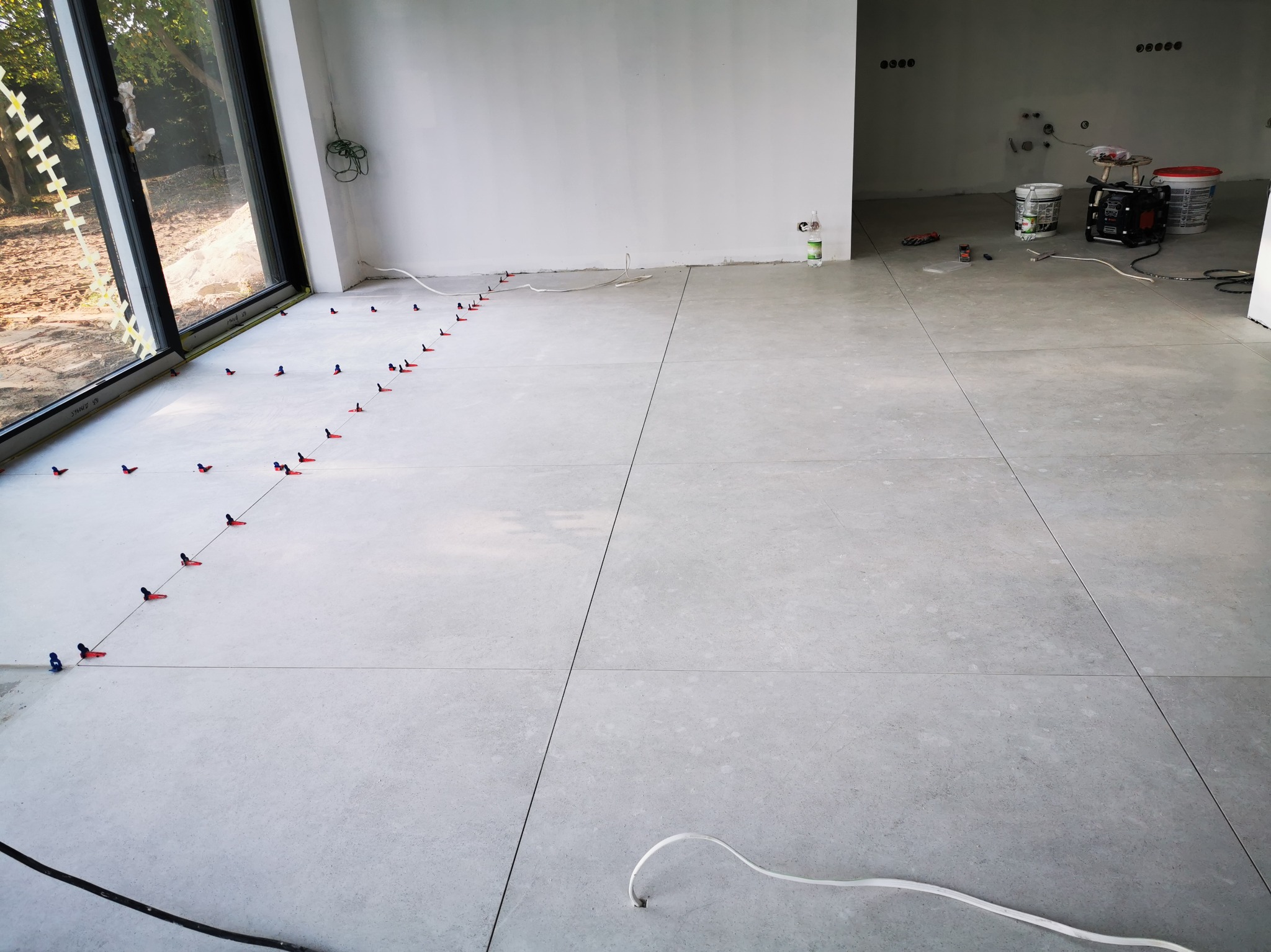 Jasnoszara podłoga z płytek w trakcie układania, z systemem poziomowania na krawędziach przy oknie i widocznymi narzędziami oraz wiadrami w tle.