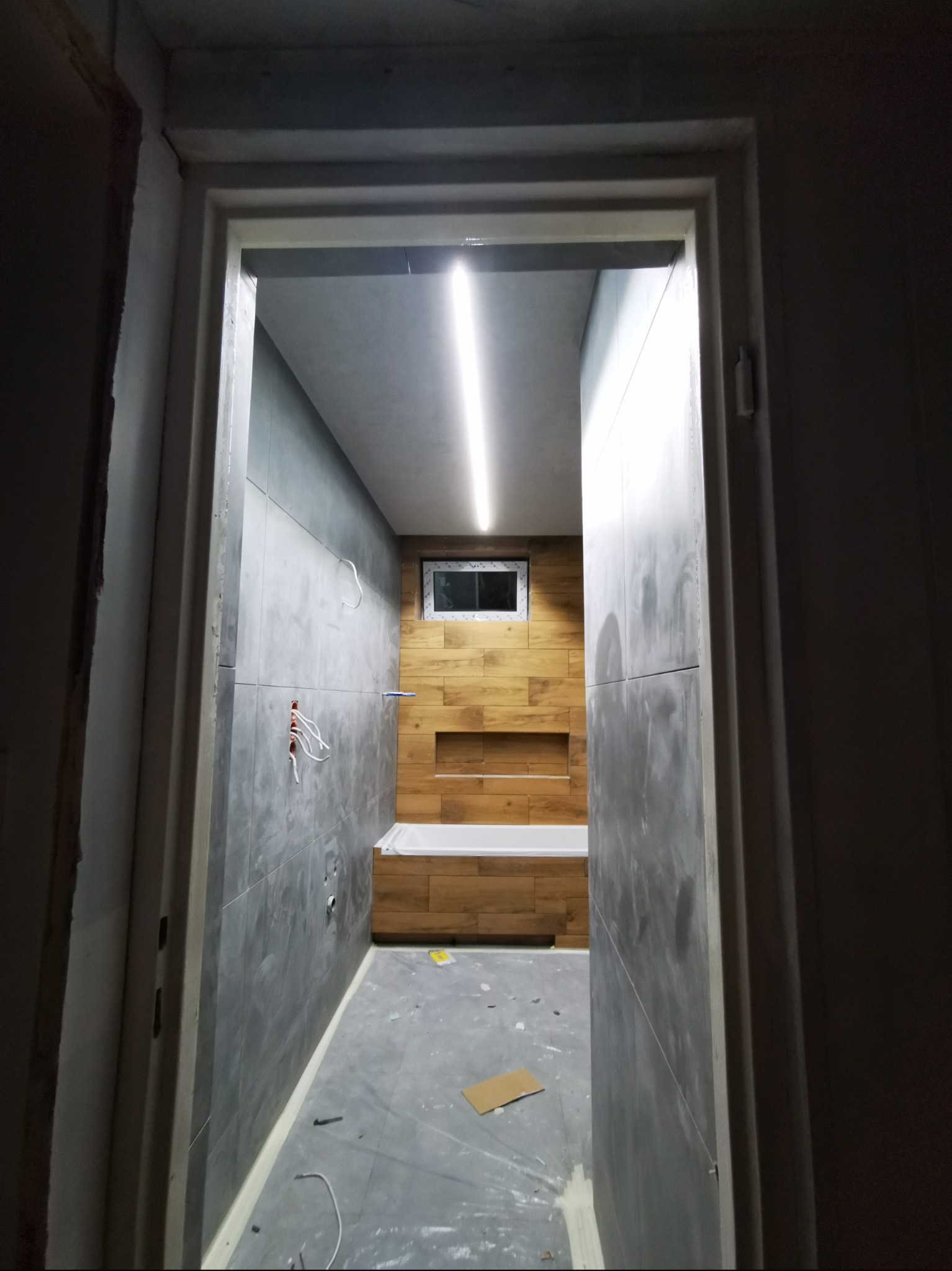 Widok przez futrynę drzwi na łazienkę w trakcie remontu: szare płytki na ścianie z instalacją elektryczną, drewnopodobne płytki wokół wanny i wnęki, surowy beton na suficie z lampą LED.