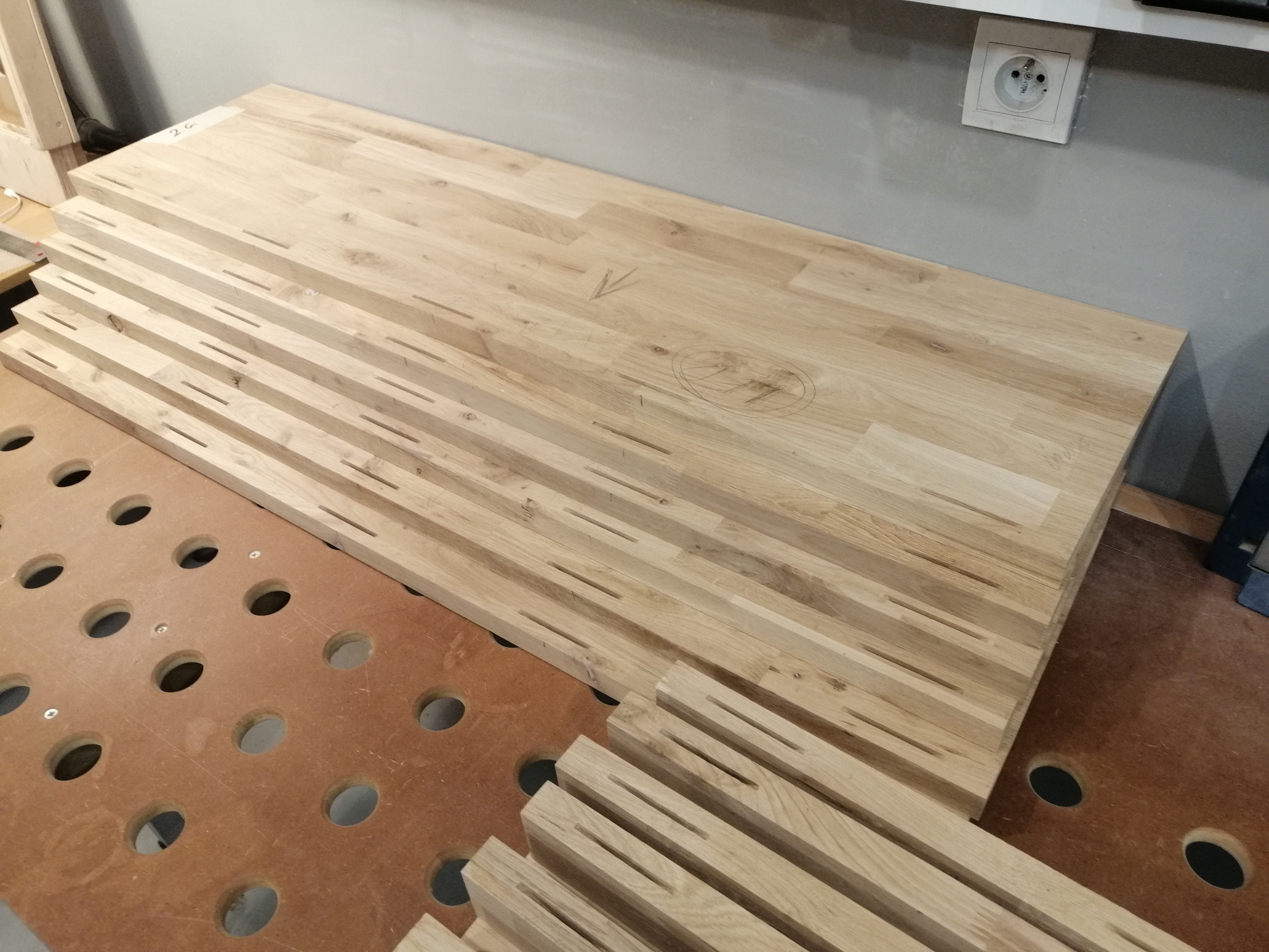 Przygotowane elementy drewniane z wpustami do łączenia, prawdopodobnie na blat, leżące na stole warsztatowym z otworami, widoczne oznaczenia ołówkiem na powierzchni.