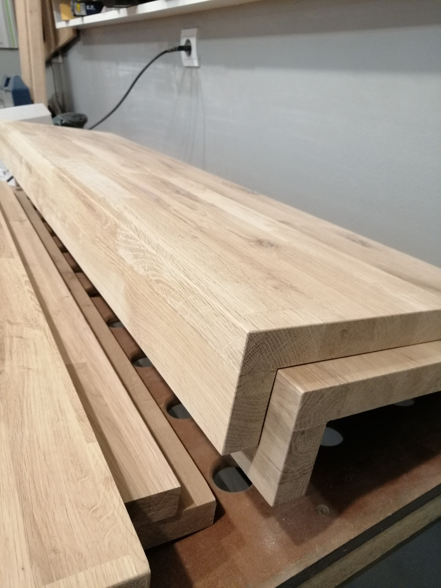 Surowe, jasne blaty drewniane z widocznym usłojeniem, jeden z nich z charakterystycznym wycięciem, ułożone na drewnianej konstrukcji w warsztacie.