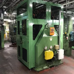 Zielona, kanciasta obudowa maszyny przemysłowej z kratkami wentylacyjnymi i pomarańczową skrzynką kontrolną z przyciskami, widoczna w hali produkcyjnej z rurami i przewodami na suficie