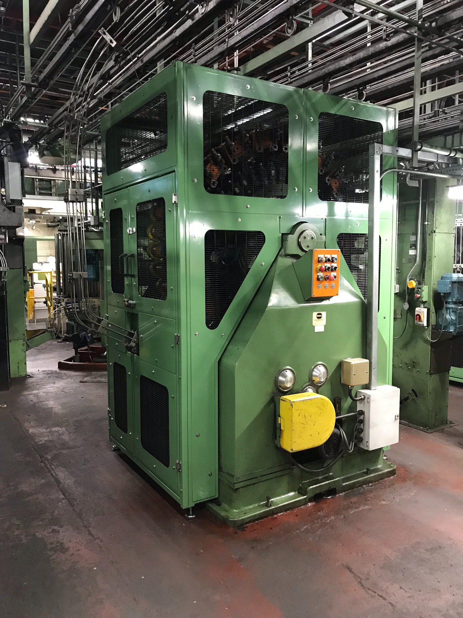 Zielona, kanciasta obudowa maszyny przemysłowej z kratkami wentylacyjnymi i pomarańczową skrzynką kontrolną z przyciskami, widoczna w hali produkcyjnej z rurami i przewodami na suficie