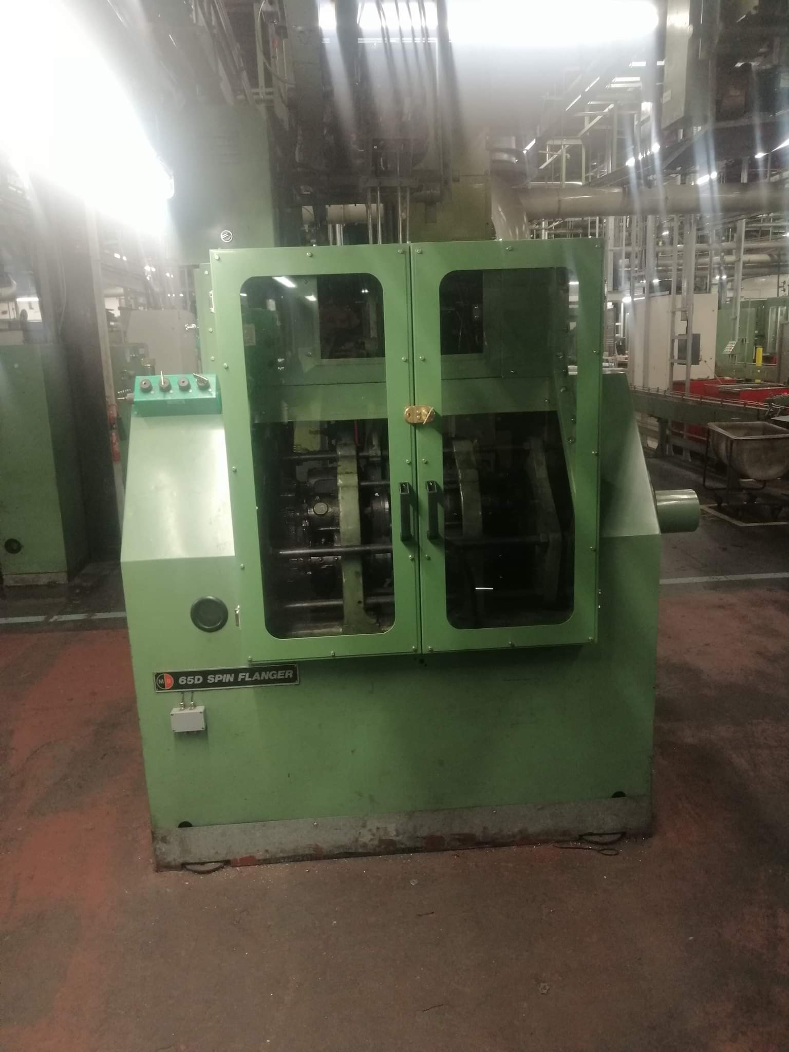 Zielona maszyna przemysłowa z osłoną z pleksi i panelem sterowania, na tle hali produkcyjnej z instalacjami rurowymi