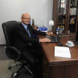 Mężczyzna w garniturze siedzi przy biurku z laptopem, obok filiżanka, otwarty kodeks i statuetka Temidy