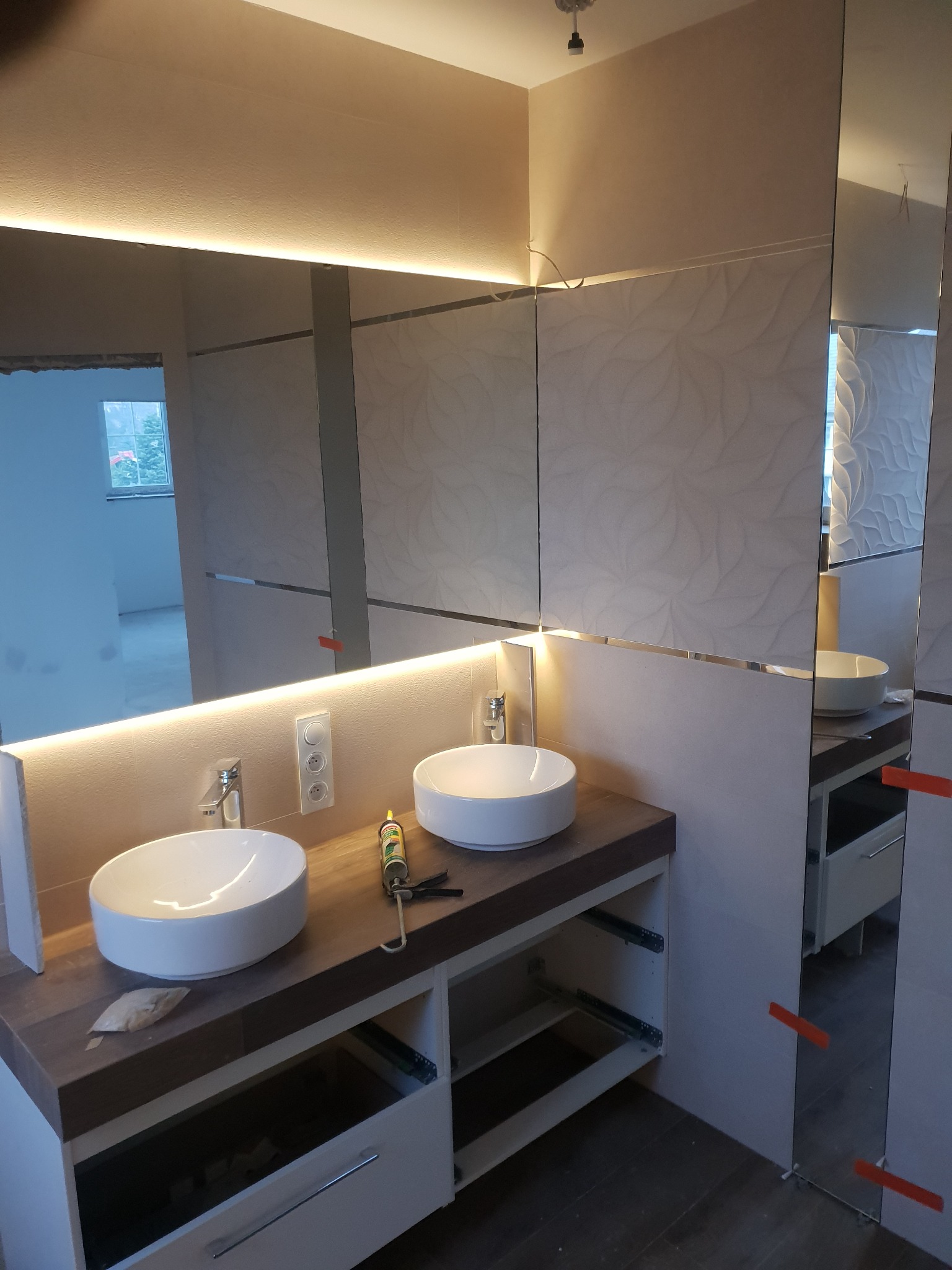 Dwie umywalki nablatowe na drewnianym blacie z szafkami w trakcie montażu, widoczne narzędzia i materiały, podświetlane lustro z efektem dekoracyjnej ściany w tle, pomieszczenie w trakcie prac...