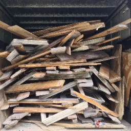 Załadunek drewna i elementów stolarki okiennej do wnętrza kontenera transportowego, widoczne fragmenty ogrodu i drzew za kontenerem.