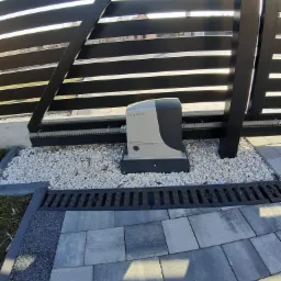 Zamontowany napęd do bramy przesuwnej Nice Robus, widoczny mechanizm z zębatką, podsypany białym grysem, z odpływem liniowym i kostką brukową w tle.