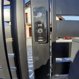 Czarny wideodomofon firmy Zamel zamontowany na nowoczesnej, metalowej bramie wjazdowej, widok na dom z brązowymi drzwiami i częściowo widoczny czerwony samochód w tle.