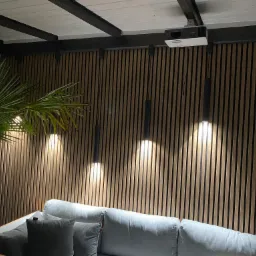Nowoczesny salon na zewnątrz z drewnianą ścianą z lameli, trzema wiszącymi lampami, projektorem sufitowym i palmą w donicy.