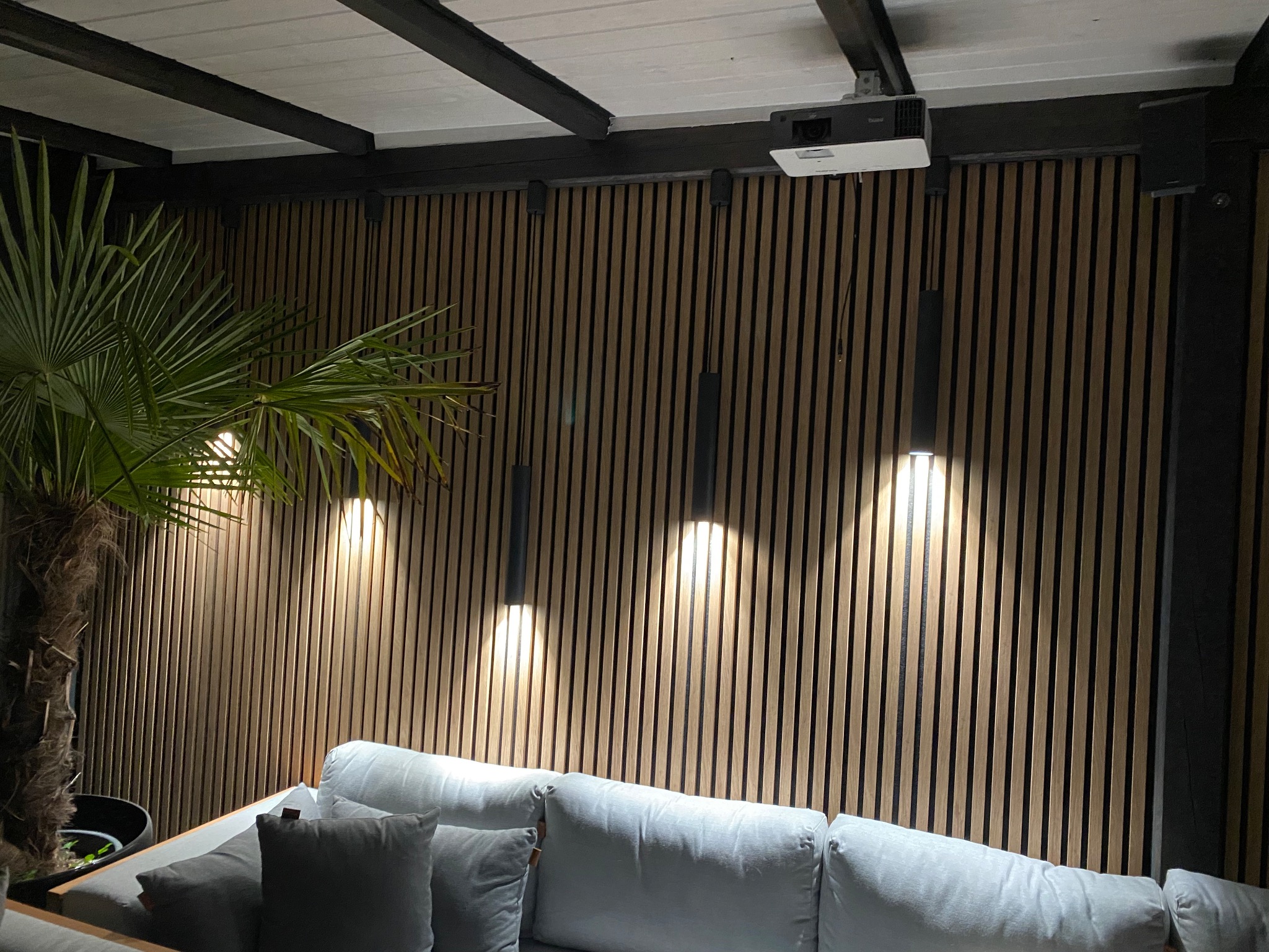 Nowoczesny salon na zewnątrz z drewnianą ścianą z lameli, trzema wiszącymi lampami, projektorem sufitowym i palmą w donicy.