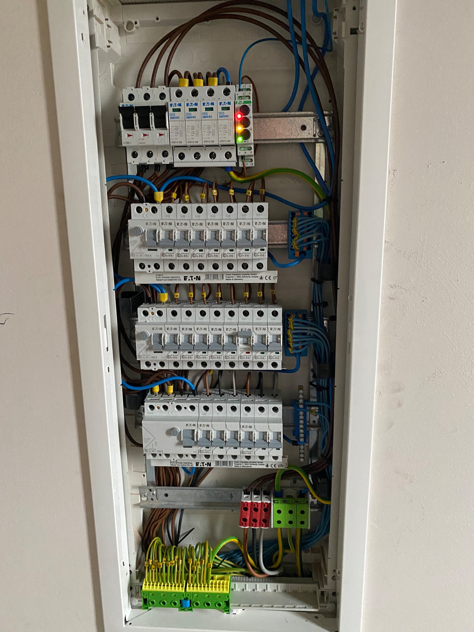 Otwarta skrzynka elektryczna z widocznymi bezpiecznikami, przewodami i listwami zaciskowymi marki Eaton, z diodami sygnalizacyjnymi w górnej części, zamontowana na jasnej ścianie.