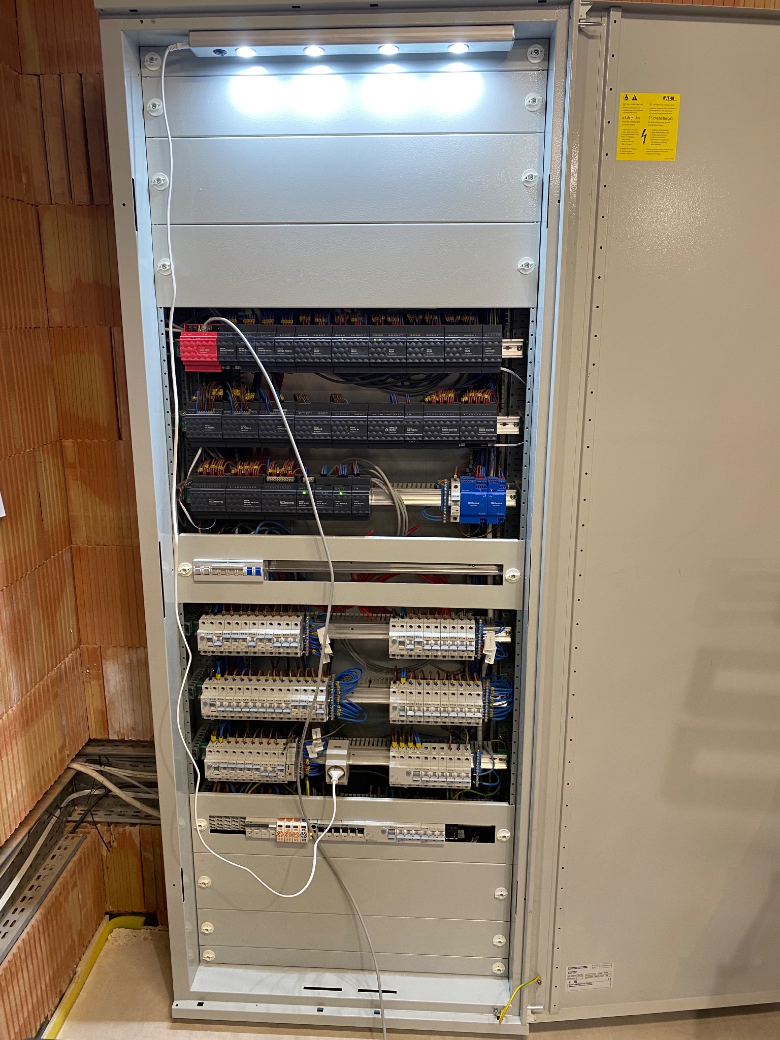 Otwarta szafa sterownicza z widocznymi modułami, bezpiecznikami i okablowaniem, oświetlona od góry, z fragmentem ściany z cegły dziurawki w tle.
