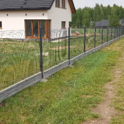 Nowoczesne ogrodzenie panelowe z podmurówką betonową otacza dom z ciemnym dachem na tle zieleni.