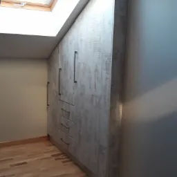 Biała szafa z szarym wzorem, dopasowana do skosu dachu, z długimi, pionowymi uchwytami i trzema szufladami na dole, na tle drewnianej podłogi i okna dachowego.