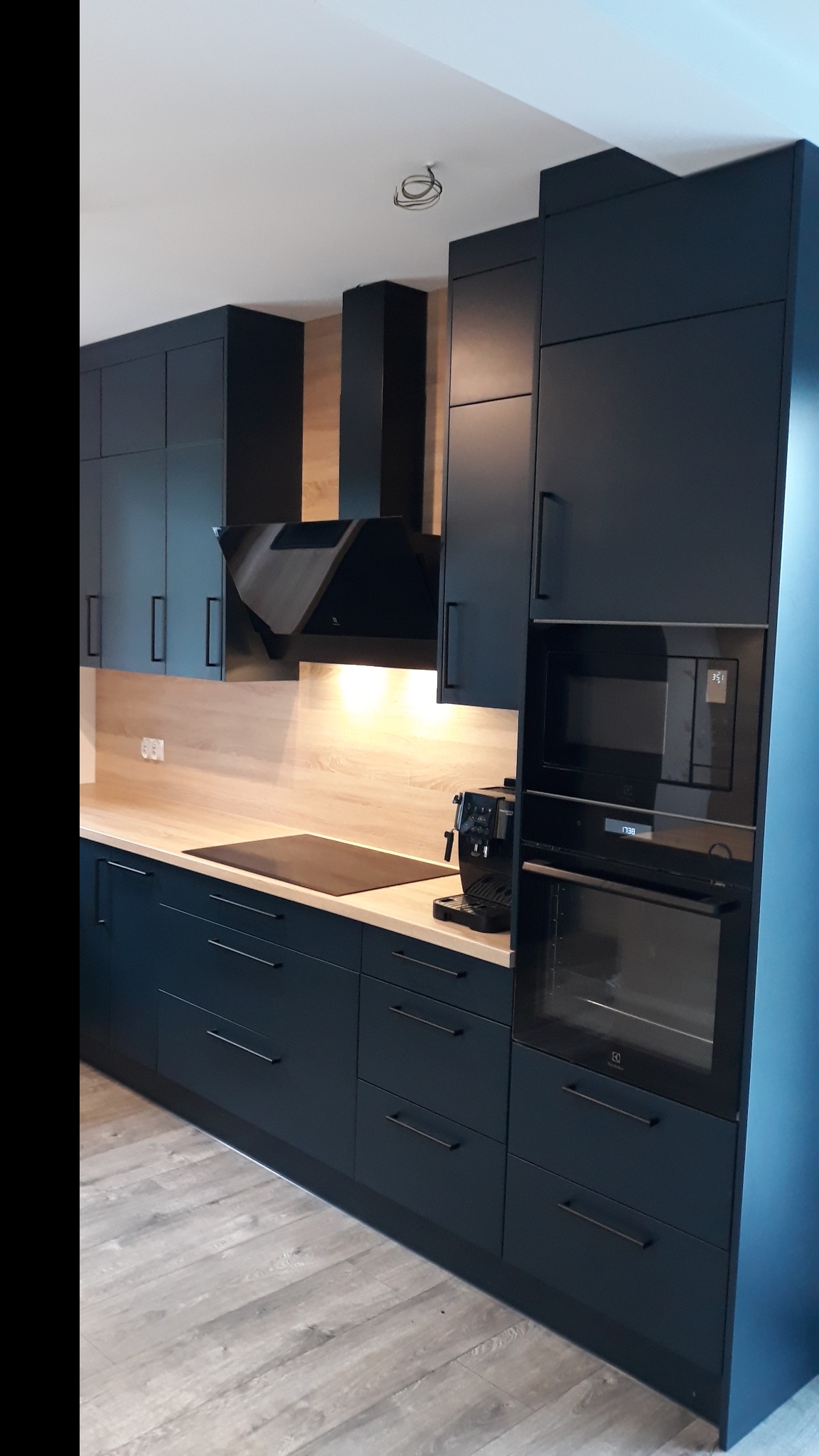 Nowoczesna, ciemnogranatowa kuchnia z drewnianym blatem i sprzętem AGD w zabudowie. Minimalistyczny design z długimi, prostymi uchwytami.