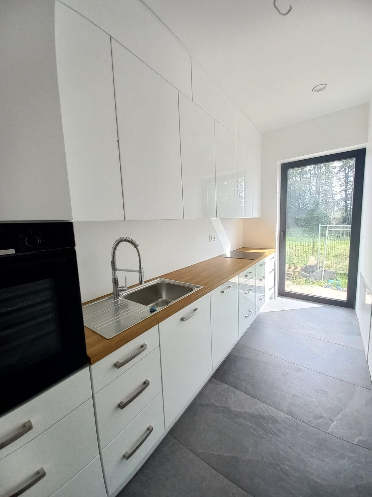 Biała kuchnia na wymiar z drewnianym blatem i szarą podłogą. Widok na zewnątrz przez czarne drzwi balkonowe. Minimalistyczny design, nowoczesne wykończenie.