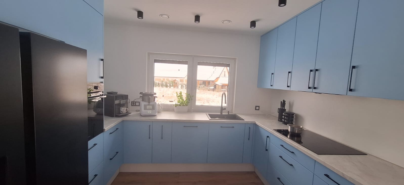 Jasna kuchnia na wymiar z błękitnymi frontami i czarnymi uchwytami. Na blacie ekspres do kawy i robot kuchenny. Widok na ogród przez okno.
