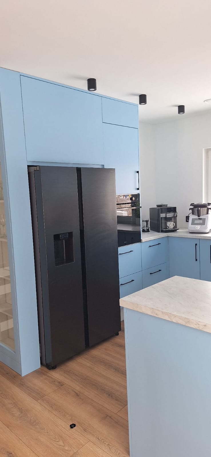 Błękitna kuchnia z czarną lodówką side-by-side, wyspą i sprzętami AGD. Minimalistyczne oświetlenie punktowe. Nowoczesny design.