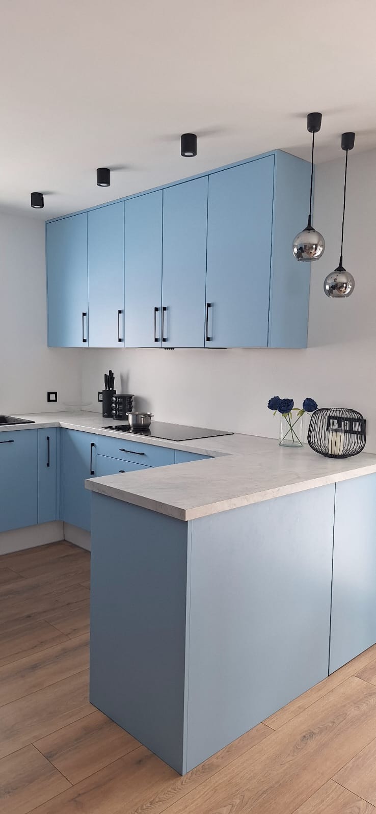 Błękitna kuchnia na wymiar z czarnymi uchwytami i lampami. Blat w odcieniach szarości, drewniana podłoga. Minimalistyczny design z dekoracjami.