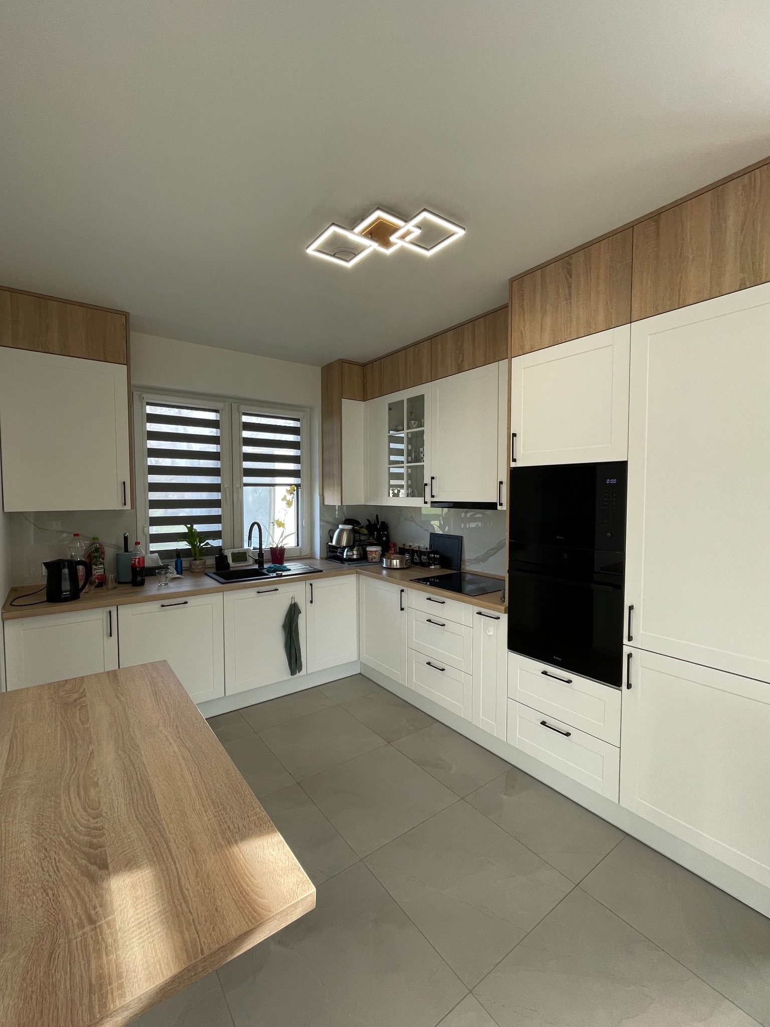 Jasna kuchnia z białymi szafkami i drewnianymi akcentami, nowoczesne oświetlenie sufitowe, szare płytki podłogowe i drewniany stół w słonecznym świetle.