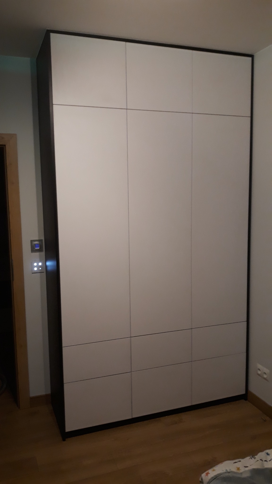 Wysoka szafa wnękowa z białymi frontami i ciemnymi obramowaniami, podzielona na sześć paneli, wkomponowana w jasne wnętrze z drewnianą podłogą.