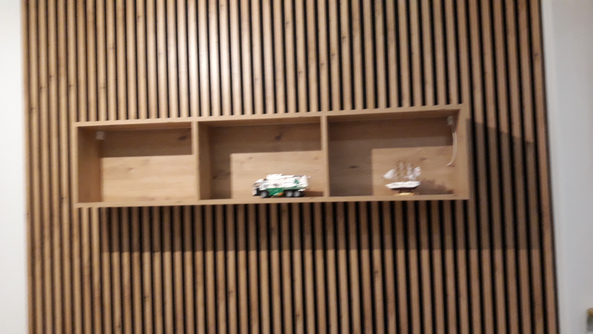 Drewniana półka z trzema przegródkami na tle ściany z drewnianych listewek. Na półce zabawka - śmieciarka i model żaglówki.