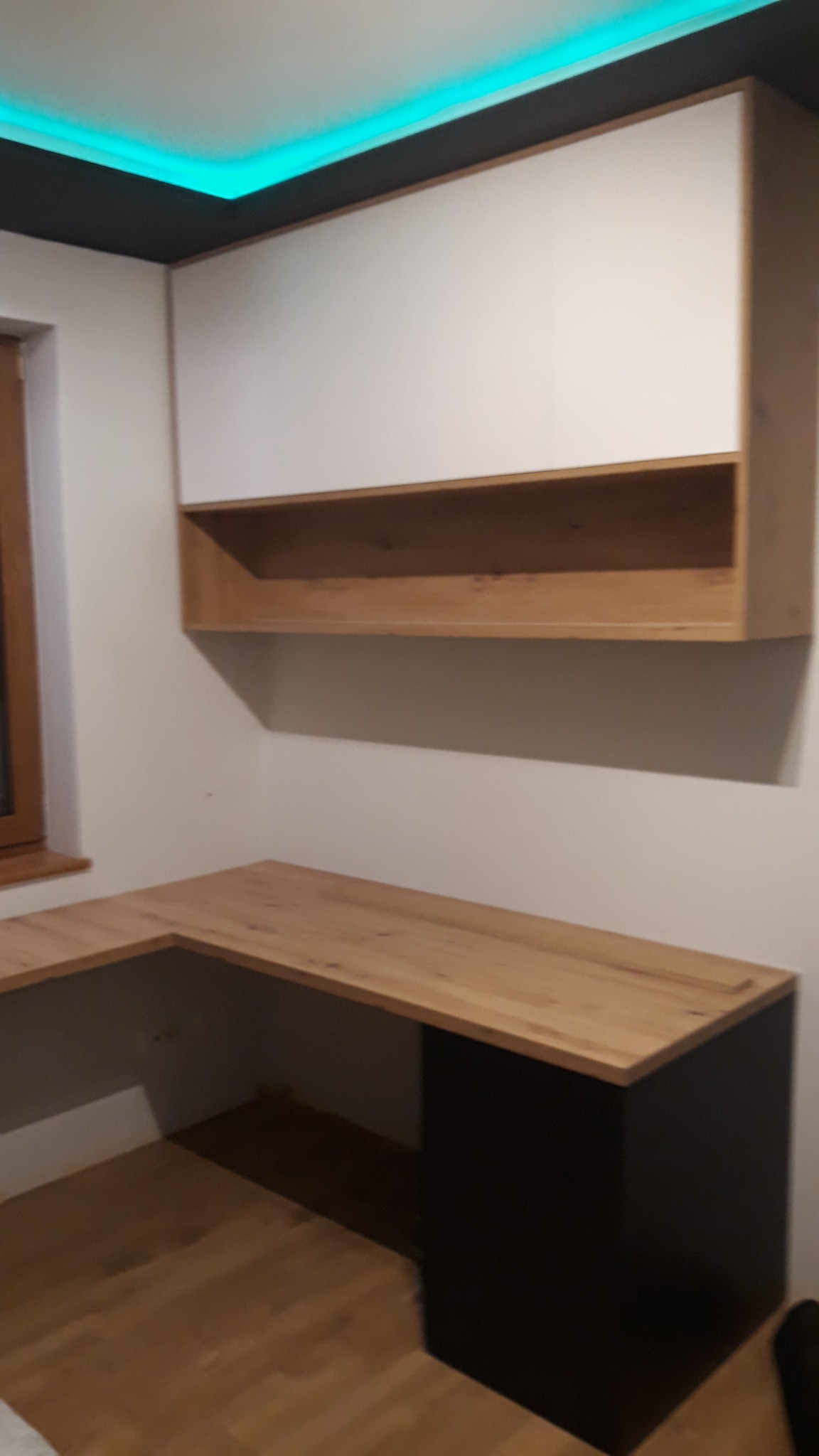 Narożne biurko z drewnianym blatem i czarną podstawą, nad nim szafka wisząca z białymi frontami i otwartą półką z drewna, oświetlenie LED na suficie.