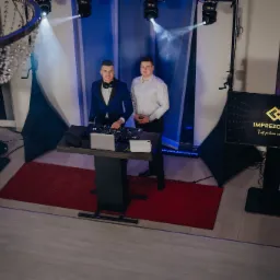 Dw&oacute;ch mężczyzn, jeden w błękitnej marynarce i muszce, drugi w białej koszuli, stoi za konsoletą DJ-ską z laptopem, na czerwonym dywanie, z oświetleniem punktowym i ekranami z logo firmy w tle.