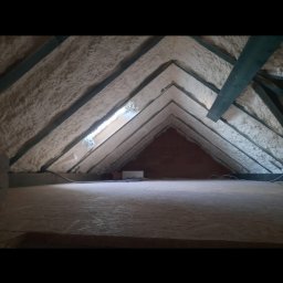 Eko Styl PUR Michał Horeczy - Poddasze z widoczną izolacją pianką PUR na skośnych powierzchniach dachu, drewniane elementy konstrukcyjne, w tle mur z cegły. Widok z poziomu podłogi.