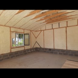 Eko Styl PUR Michał Horeczy - Wnętrze budynku w trakcie prac izolacyjnych. Widoczna pianka izolacyjna pokrywająca ściany i sufit, drewniana konstrukcja oraz okno z widokiem na zieleń.