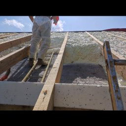 Eko Styl PUR Michał Horeczy - Aplikacja pianki izolacyjnej na poddaszu, widok z dołu na krokwie i pracownika na drabinie, gotowa warstwa piany PUR, jasne niebo w tle.