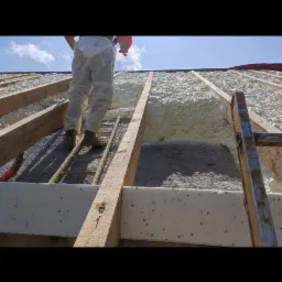 Aplikacja pianki izolacyjnej na poddaszu, widok z dołu na krokwie i pracownika na drabinie, gotowa warstwa piany PUR, jasne niebo w tle.