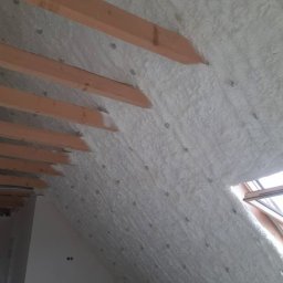 Eko Styl PUR Michał Horeczy - Poddasze po natrysku pianki izolacyjnej, widoczne drewniane krokwie i biała, równomierna warstwa izolacji, okno dachowe w tle.