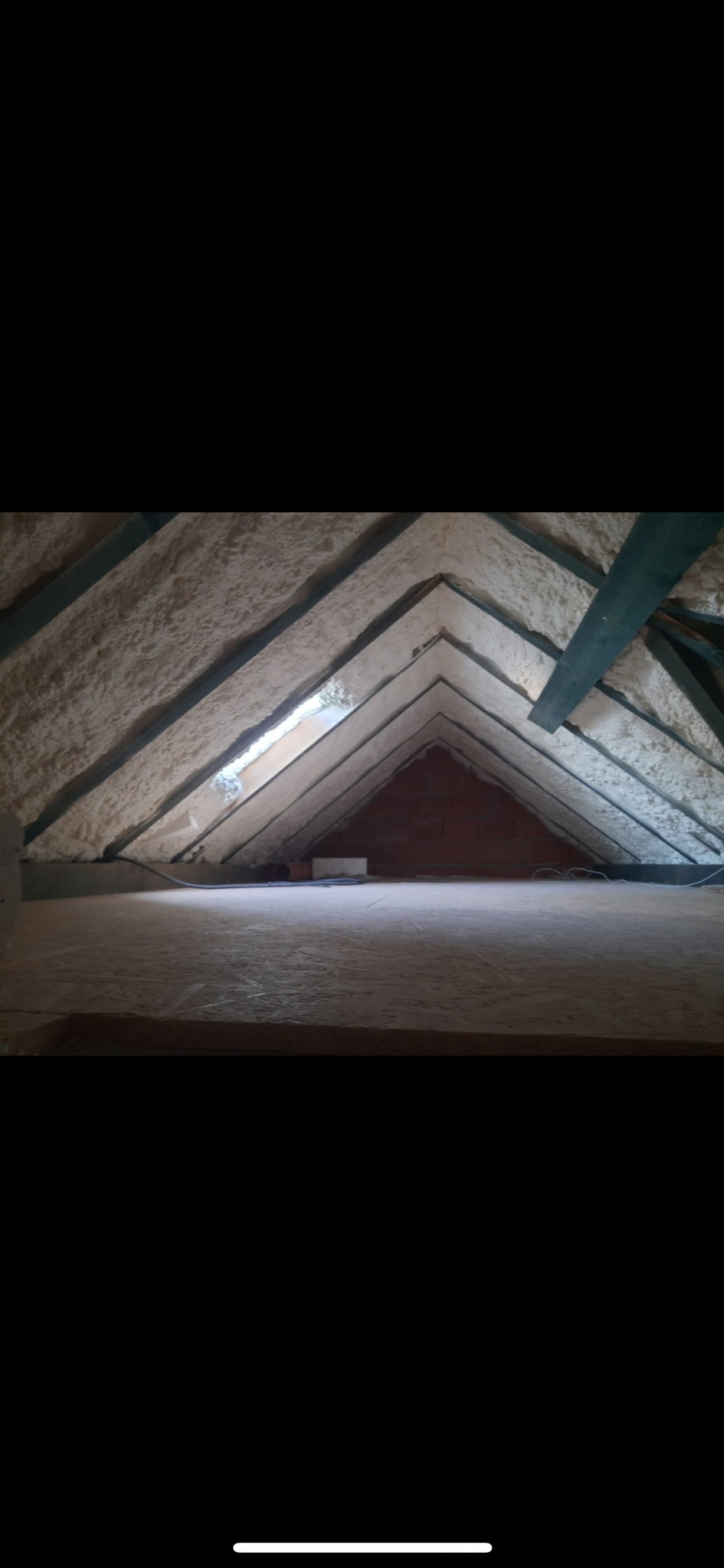 Poddasze z widoczną izolacją pianką PUR na skośnych powierzchniach dachu, drewniane elementy konstrukcyjne, w tle mur z cegły. Widok z poziomu podłogi.