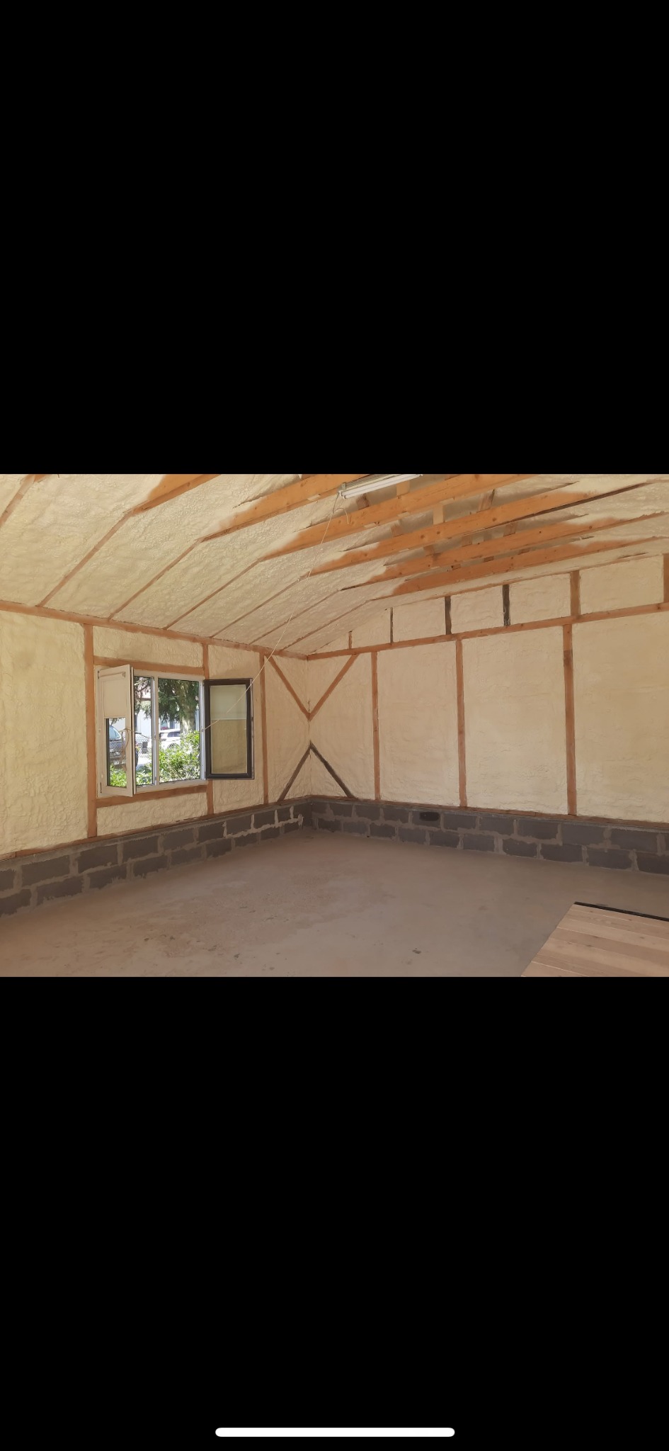 Wnętrze budynku w trakcie prac izolacyjnych. Widoczna pianka izolacyjna pokrywająca ściany i sufit, drewniana konstrukcja oraz okno z widokiem na zieleń.