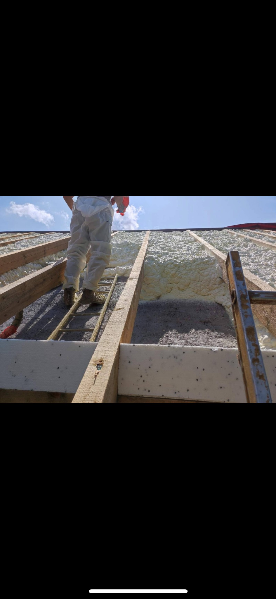 Aplikacja pianki izolacyjnej na poddaszu, widok z dołu na krokwie i pracownika na drabinie, gotowa warstwa piany PUR, jasne niebo w tle.