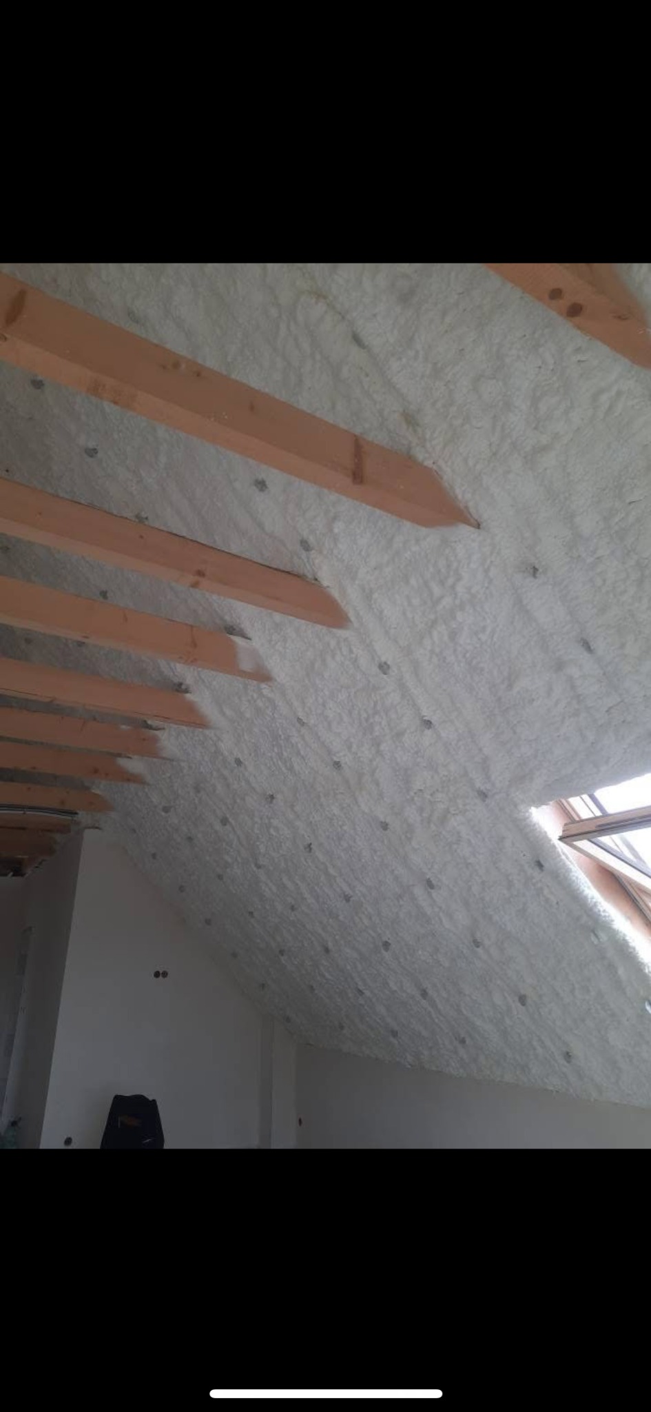 Poddasze po natrysku pianki izolacyjnej, widoczne drewniane krokwie i biała, równomierna warstwa izolacji, okno dachowe w tle.