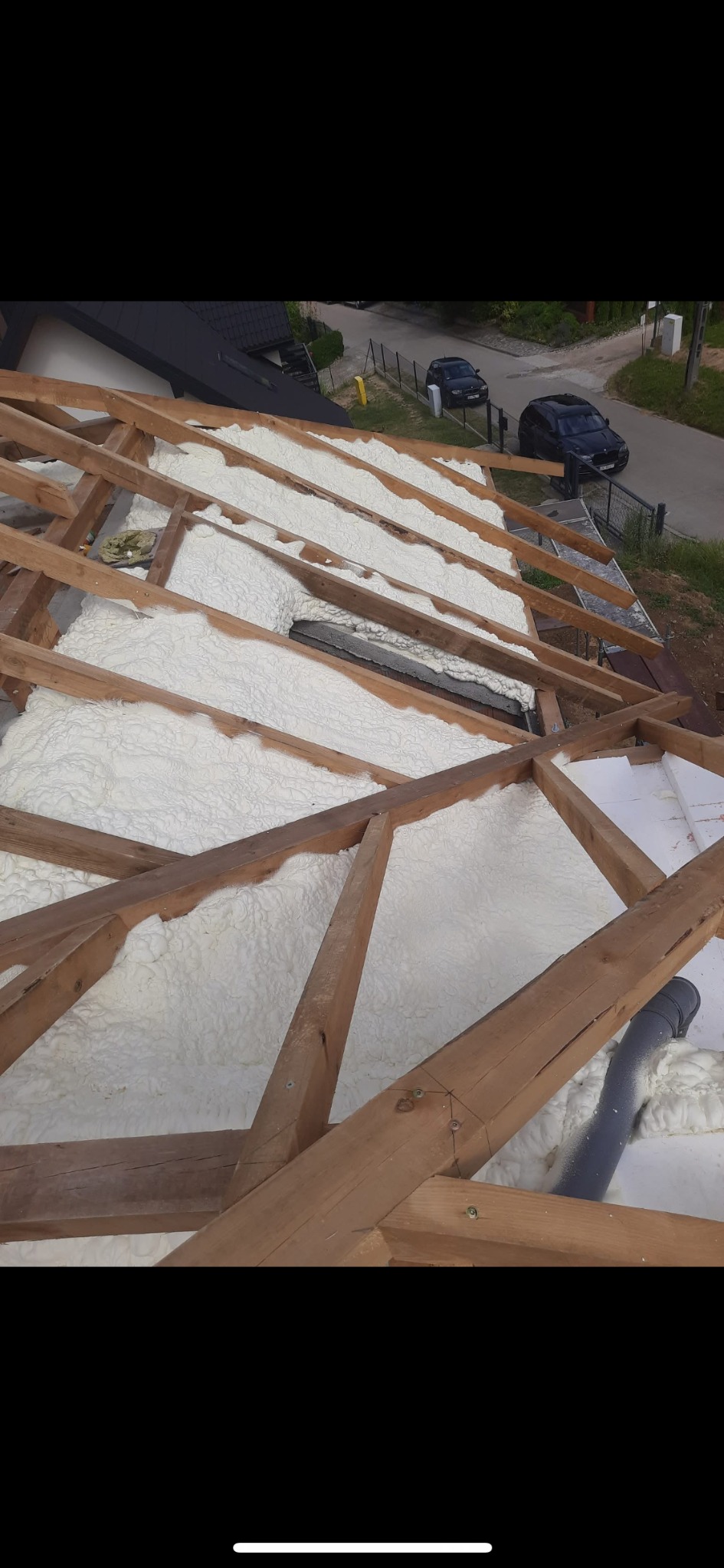 Ocieplenie poddasza pianką PUR, widok na drewnianą konstrukcję dachu wypełnioną białą pianą izolacyjną. W tle zabudowa jednorodzinna i zaparkowane samochody.