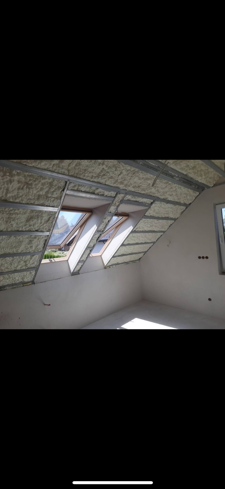 Poddasze z oknami dachowymi po aplikacji pianki izolacyjnej między metalowymi profilami, widok na fragment wnętrza, w tle zieleń za oknem.