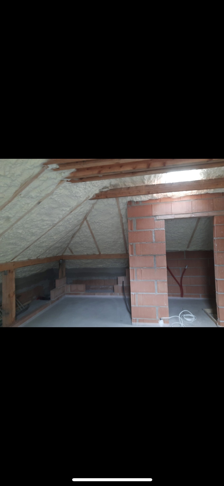 Poddasze z drewnianą konstrukcją po ociepleniu pianką: jasna izolacja pokrywa skośne ściany, widoczne ceglane elementy i fragment instalacji elektrycznej.