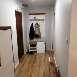 Przedpokój w odcieniach szarości z drewnianą podłogą i meblami, widoczne wieszaki z ubraniami, szafka na buty i oświetlenie sufitowe.