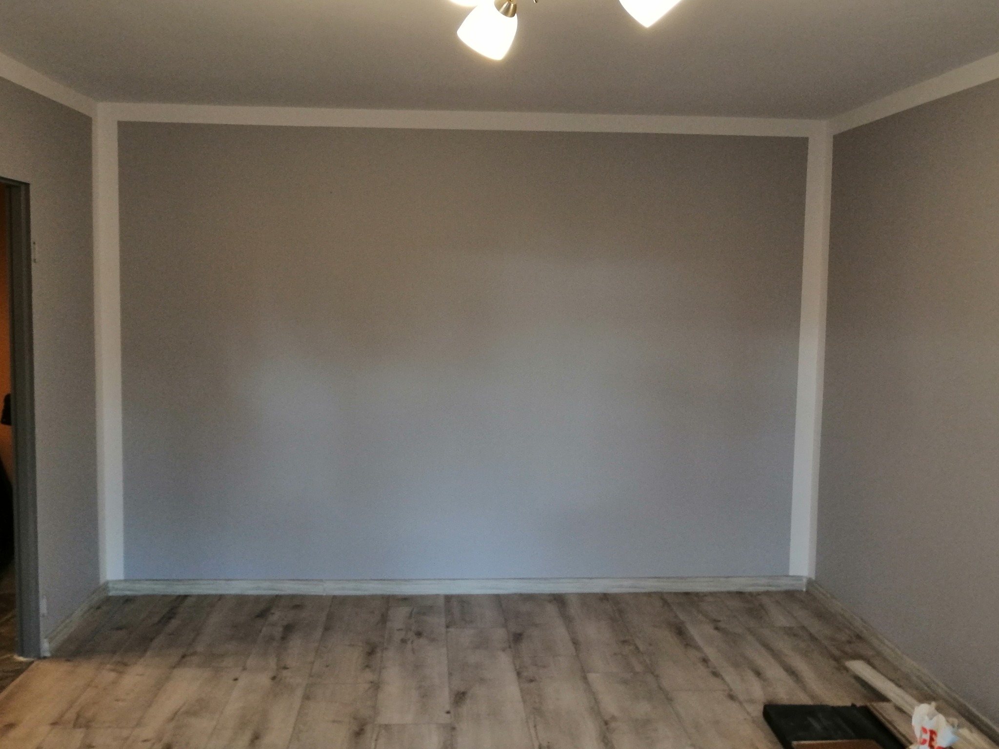 Szare ściany z białym obramowaniem i panelami podłogowymi w odcieniach drewna, widoczne narzędzia do układania paneli.