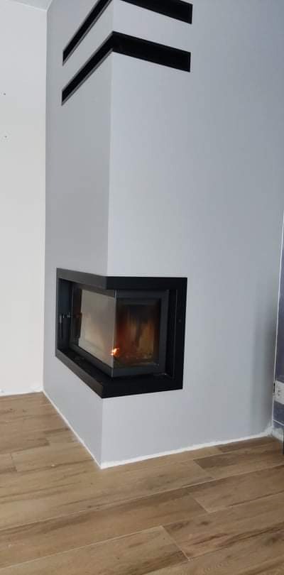 Wnętrze z narożnym kominkiem obudowanym w szarej ścianie z czarnymi, poziomymi dekoracjami, na podłodze panele imitujące drewno.